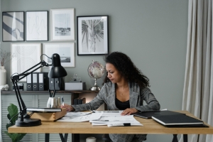 A woman working at a desk in a home office, surrounded by documents and framed artwork on the wall.