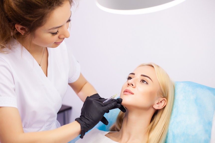 A cosmetic practitioner wearing black gloves is administering an injection to a woman’s chin as she lies on a treatment chair in a clinical setting.
